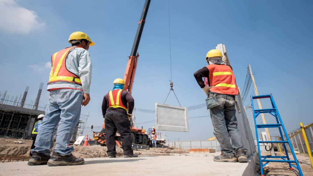 3 homens olhando um guindaste elevando uma peça de concreto.