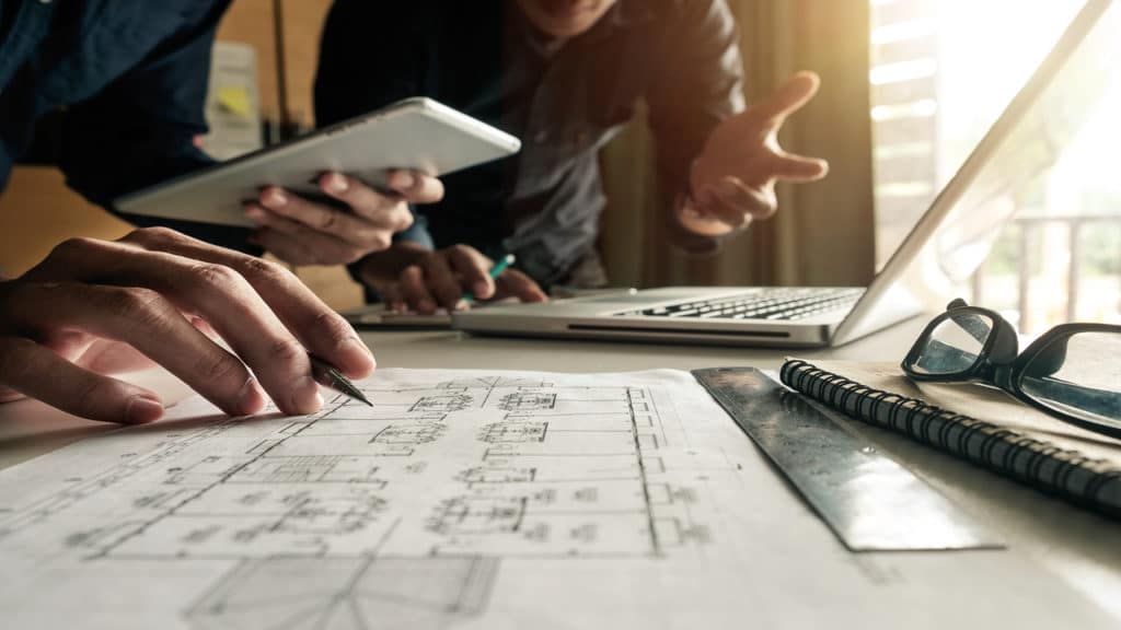 Dois homens conversando, um está a frente de um notebook e o outro está olhando a planta arquitetônica de alguma obra.