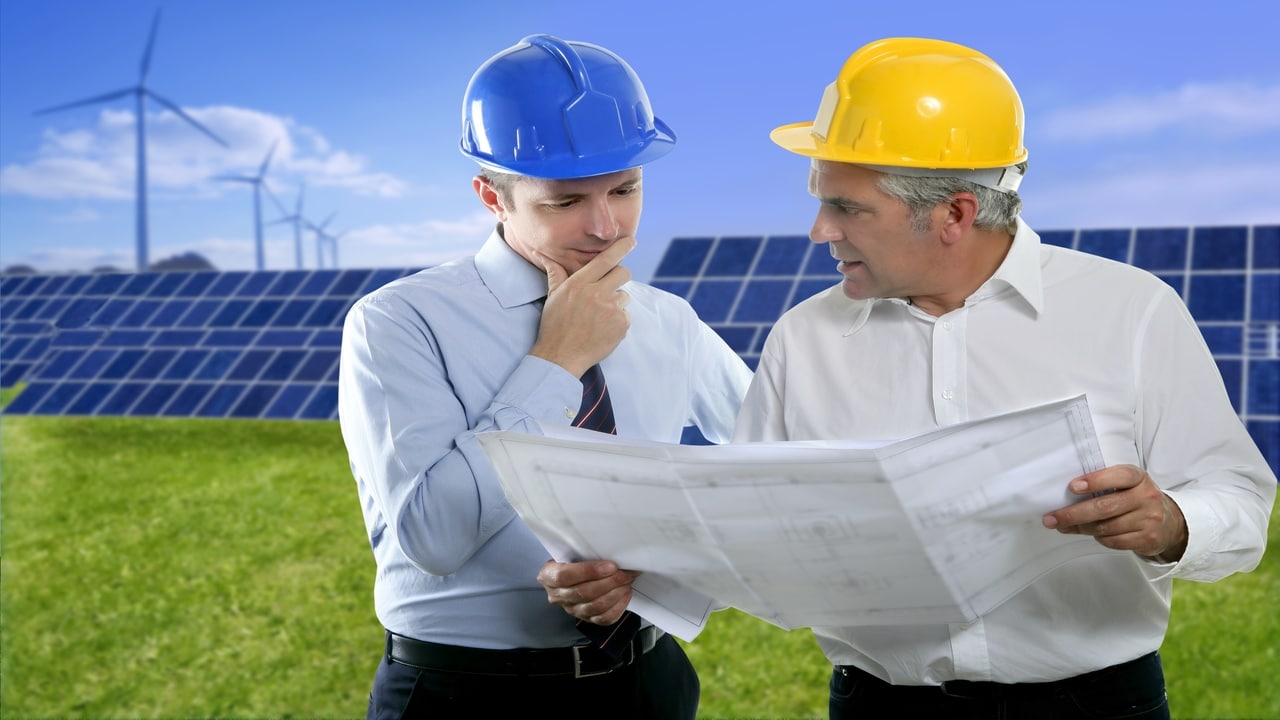 Dois homens olhando a planta de um projeto e ao fundo placas solares e hélices eólicas.