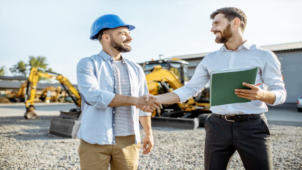 Dois homens se cumprimentando e um ambiente de obras ao fundo.