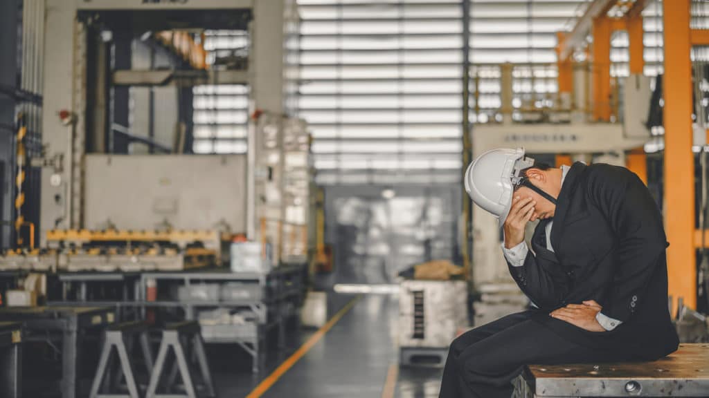 Homem com a mão em sua cabeça, mostrando tristeza, dentro de um galpão de materiais.