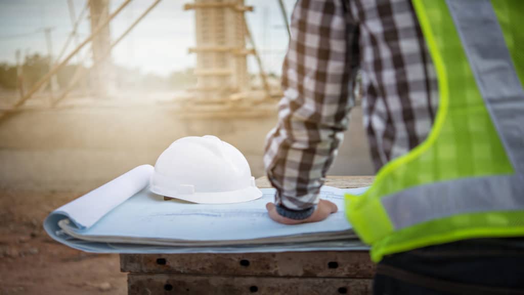 Engenheiro de costas olhando a planta de sua obra e uma paisagem de obras ao fundo.