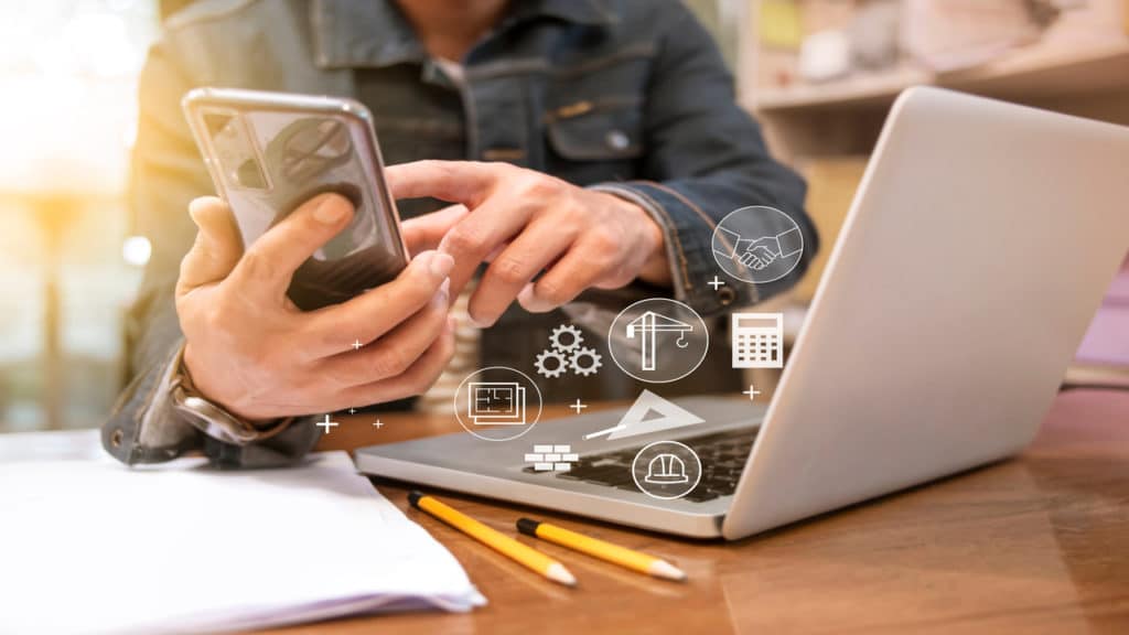 Homem olhando o celular em frente ao seu notebook, planejando suas finanças da obra.