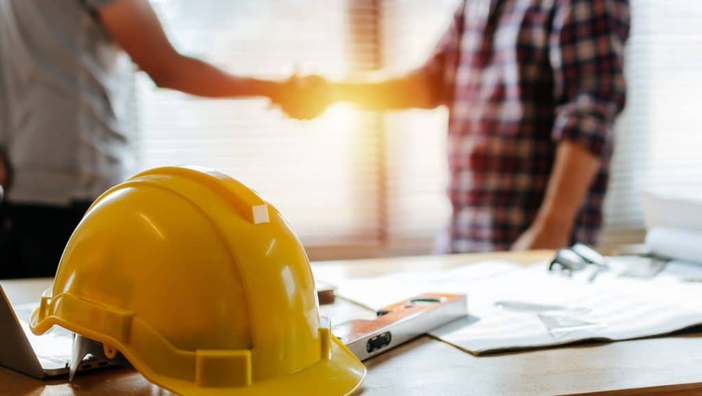 Dois homens apertando as mãos e um capacete de obra em cima da mesa