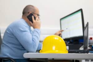 Um homem falando ao telefone enquanto olha para a tela de um computador, com um capacete de segurança amarelo sobre a sua mesa