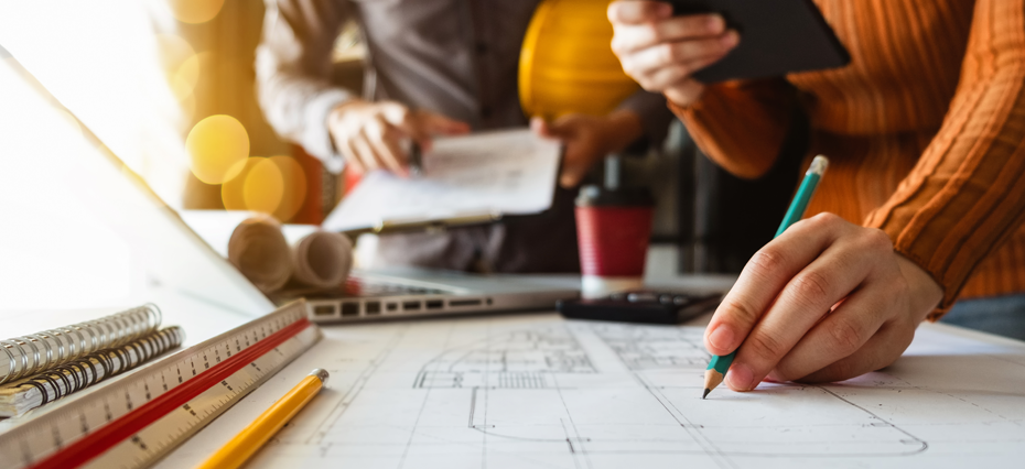 Mãos desenhando em um papel que está sobre a mesa. Há um computador ao lado do que parece ser o plano de marketing de um fornecedor que pretende anunciar para construtoras